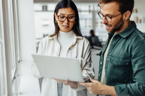 Business professionals standing in an office and using a laptop Business professionals standing in an office and using a laptop together. Two young business people having a discussion in a workplace. casual clothing stock pictures, royalty-free photos & images