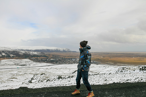 A hiker tourist man in winter clothes is standing on a mountain peak and looking at a small icelandic city, by spring