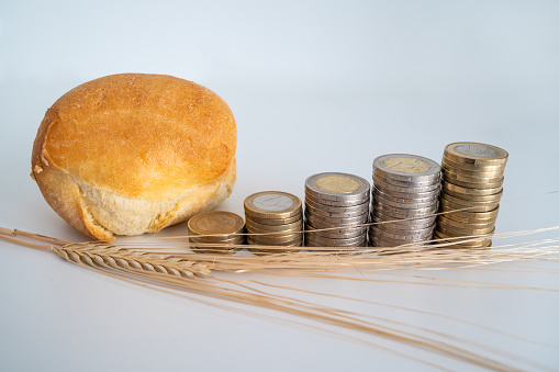 Piece of bread, next to stacks of coins of increasing height, and an ear of corn. Increase and trend in the price of wheat and bread.