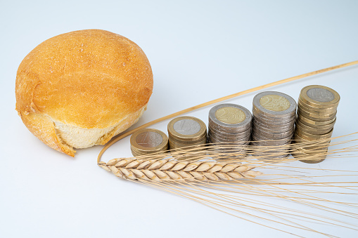 Piece of bread, next to stacks of coins of increasing height, and an ear of corn. Increase and trend in the price of wheat and bread.