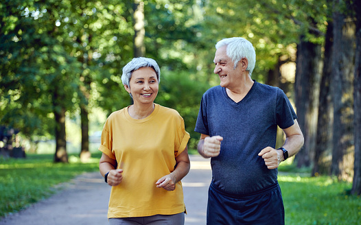 Mature smiling grey-haired fit couple jogging in the summer park. Two older joggers enjoy morning active cardio outdoors, keep healthy active lifestyle. Weightloss, sport concept
