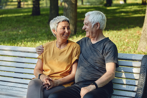 Happy multi racial elderly grey hair couple resting sit on bench in park after stroll, morning walk smiling enjoy relaxation outdoors at sunny summer day. Fit family, break, love concept
