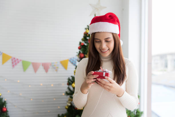 uśmiechnięta brunetka w swetrze i czapce świętego mikołaja trzyma małe pudełko prezentowe i cieszy się z bożego narodzenia dekoracyjnego białego tła - expressing negativity sullen 20s attractive male zdjęcia i obrazy z banku zdjęć