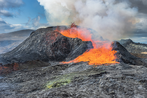 Lava fountains from the volcanic crater in Iceland. Landscape on Reykjanes peninsula in spring with sunshine. Liquid lava flows out of the side of the crater. Strong steam rises from crater