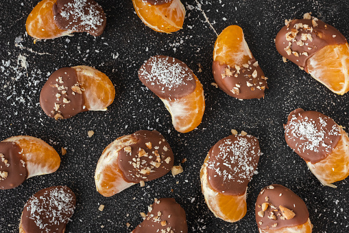 Orange juicy tangerine slices covered with melted milk chocolate on black concrete background. Sweet appetizer