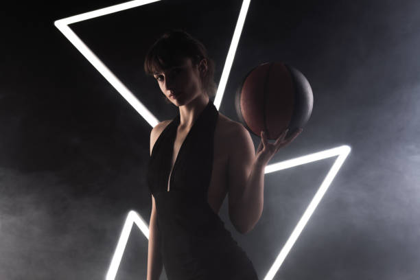 Beautiful girl in a fancy black dress holding basketball. Studio portrait against fog background with white neon lights. stock photo