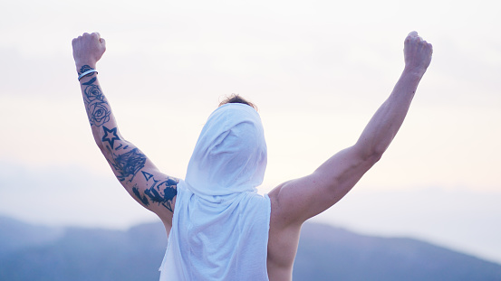 Man in a white hood standing on top of a tall mountain looking at the view below and taking it in. Enjoying the wind and celebrating success