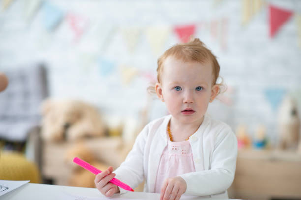 A sweet little 2 year old girl sits at a table in her daycare classroom as she colors away.  She is dressed casually and has a neutral expression on her face.