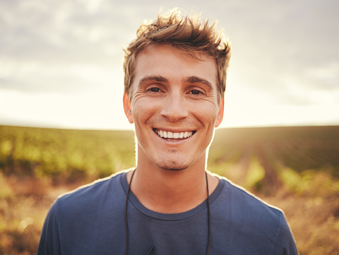 Happy, face and nature with a man at sunset, enjoying freedom with a smile and standing on a field of grass in the day. Travel, vacation and morning with the portrait of a handsome young male outside