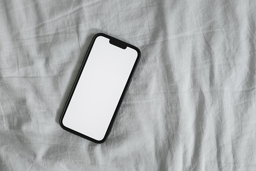 Smart mobile phone with blank mockup screen on wrinkled bed linen, top view
