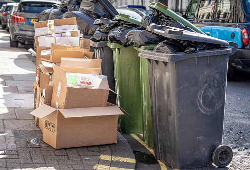 A pile of cardboard on a London street for recycling, next to a group of overflowing plastic wheel bins.