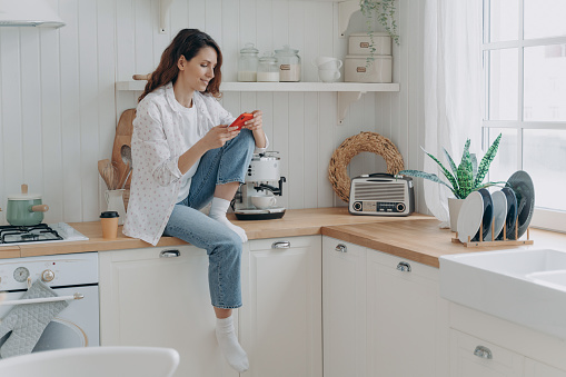Female uses modern phone, messaging online at kitchen at home, rests after chores. Woman housewife holding smartphone, shopping on internet, ordering groceries online, using delivery service app.