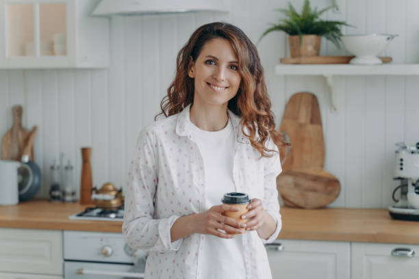 lächelnde hispanische frau mit pappbecher kaffee steht in gemütlicher moderner küche und schaut in die kamera - stereotypical housewife women domestic kitchen brown hair stock-fotos und bilder