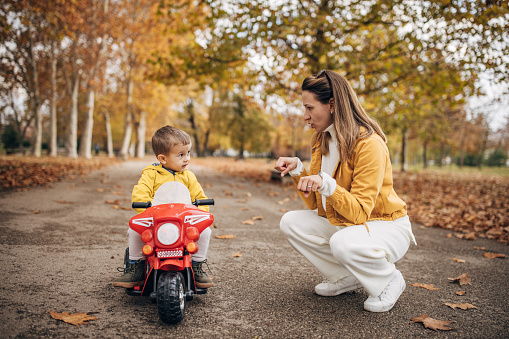 Two people, mother teaching her little son to ride his toy motorcycle on autumn day in park.