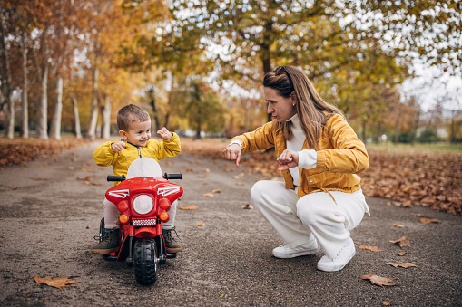 Two people, mother teaching her little son to ride his toy motorcycle on autumn day in park.