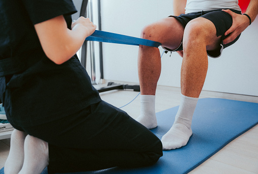 Side view of young female physiotherapist performing recovery exercises with rubber band on young man's knee in rehabilitation center.