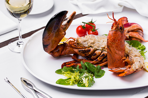 Oven baked lobster half with cheese and cream sauce. Nearby lies a sprig of cherry tomatoes and a mix of arugula and lettuce salad. The food is on a white: round plate. The plate is on a white tablecloth, next to it is a glass of white wine and cutlery.