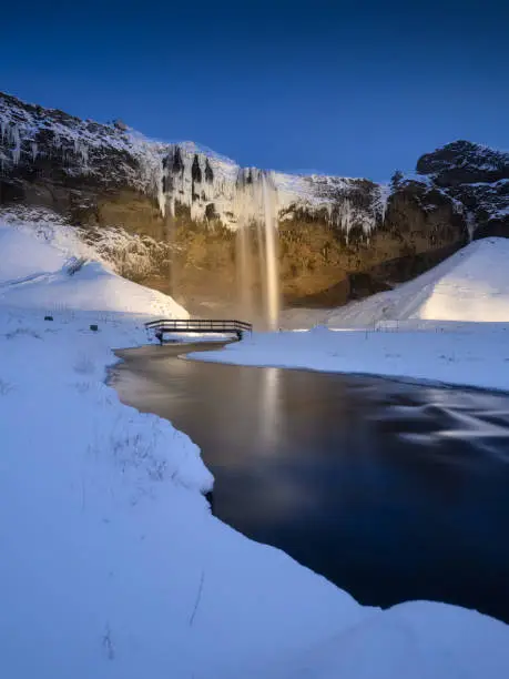 Photo of Seljalandsfoss waterfall, Iceland. Icelandic winter landscape.  High waterfall and rocks. Snow and ice. Powerful stream of water from the cliff. A popular place to travel in Iceland.