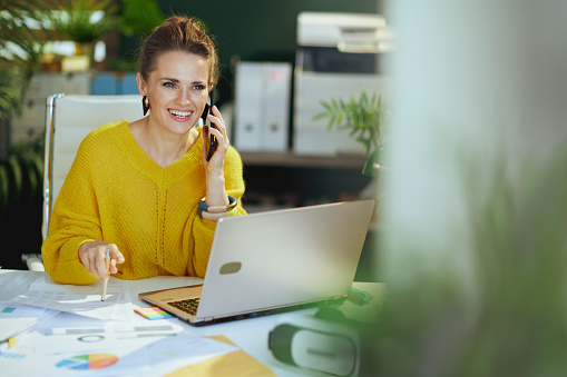 smiling trendy middle aged small business owner woman in yellow sweater with laptop speaking on a smartphone and working with documents in the modern green office.