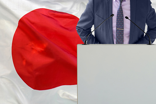 Tribune with microphone and man in suit on Japan flag background. Businessman and tribune on Japan flag background. Politician at the podium with microphones background Japan flag. Business conference
