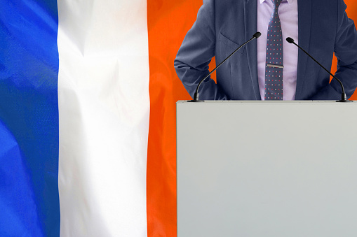 Tribune with microphone and man in suit on France flag background. Businessman and tribune on France flag background. Politician at the podium with microphones background France flag. Conference