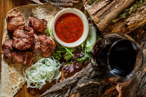 Grilled pork skewers, lying on pita bread, next to lettuce and arugula salad and sliced onion rings. A sauce-pot with adjika lies nearby, Food and sauce are on a wooden board. The board stands on a wooden background with acacia logs. A glass of red wine. brochetas de cerdo a la parrilla, acostadas sobre pan de pita, junto a ensalada de lechuga y rúcula y aros de cebolla en rodajas. una olla de salsa con adjika se encuentra cerca, la comida y la salsa están en una tabla de madera. el tablero se encuentra
