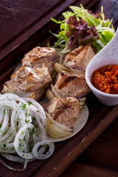 Pork skewers with grilled onions, rings of fresh onions lying side by side and strewn with parsley. Shish kebab lies on pita bread. There is a bowl with Georgian sauce nearby. Food and sauce lie on a wooden board. brochetas de cerdo con cebollas a la parrilla, aros de cebollas frescas acostados uno al lado del otro y cubiertos de perejil. shish kebab se encuentra en el pan de pita. hay un tazón con salsa georgiana cerca. la comida y la salsa se encuentran en una ta