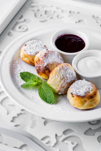 istock Cheesecakes with powdered sugar and mint on a white plate. Nearby there is a saucepan with sour cream and a saucepan with currant jam. A white plate stands on a white, wooden, carved tray. 1445220703