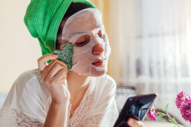 istock Woman with facial sheet mask applied using gua sha stone beauty tool at home after bath using phone. Skincare product 1445221271