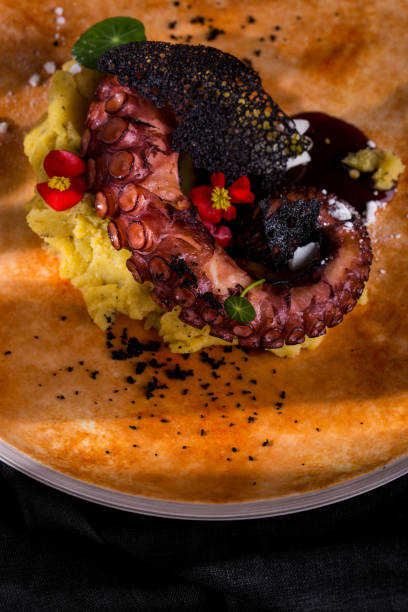 istock Fried octopus with caramel chips on mashed potatoes decorated with edible flowers. Ceramic plate on a black, linen background. 1445222548