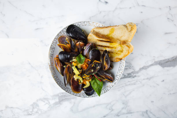 mussels fried in butter with garlic and carrots, celery and onions. on top are basil leaves and bread toast. the food lies in a deep plate with a pattern. the plate stands on a marble background. - basil tomato soup food and drink imagens e fotografias de stock