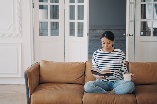 Woman is reading novel and drinks coffee or tea. Happy young european woman is reading book sitting on comfortable leather couch. Lady enjoying poetry. Living room interior with copy space.