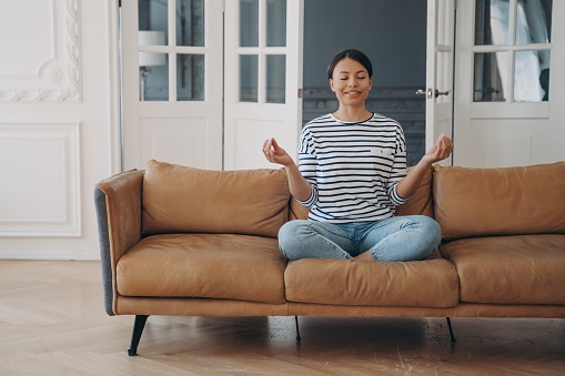 Smiling female practicing yoga, sitting in padmasana lotus pose on couch while meditation. Calm happy woman meditates with mudra gesture on sofa at home. Healthy lifestyle, wellness concept.
