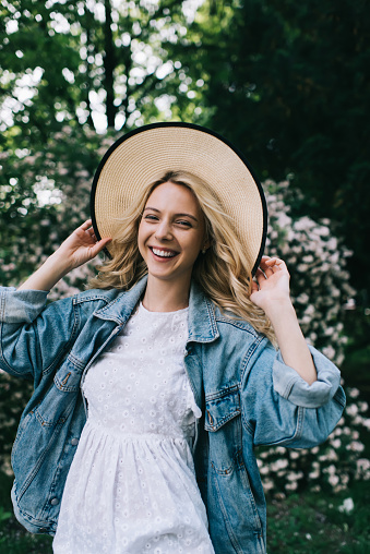 Cheerful young blonde woman in hat and jeans jacket looking at camera and laughing happily while standing in blooming park during summertime