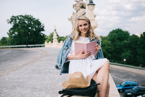 Charming young female with blond hair friendly smiling and looking at camera while sitting on border and reading book with pink cover near road