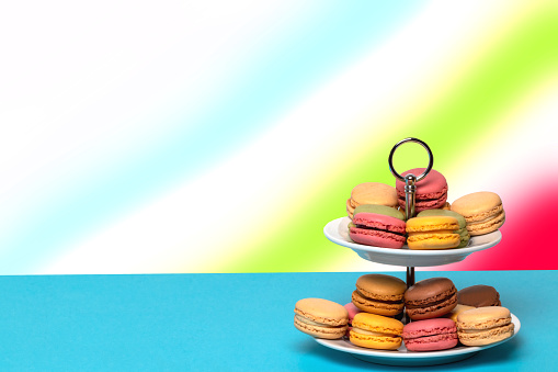 Close-up of colourful French macaroons on a two-storey etagere on a blue table with space over abstract rainbow background. Template for your food and product display montage. Pastries, desserts and sweets. Macro.