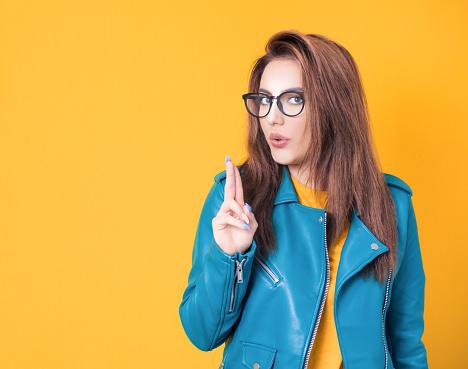 Young woman doing gun gesture getting ready to shoot, cute girl showing gun gesture, wearing blue leather jacket, isolated on yellow background. Studio shot