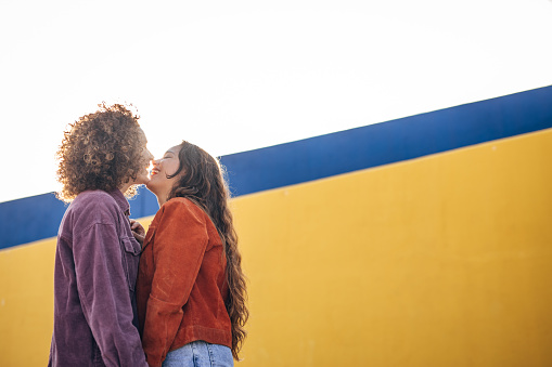 Two women, lesbian couple standing against a yellow wall outdoors. They are kissing on mouth.