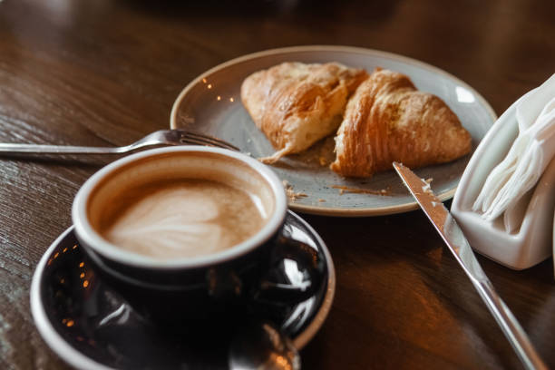 A girl eats a croissant in a restaurant and drinks latte coffee at a wooden table. Cutting a croissant on a plate, close-up stock photo