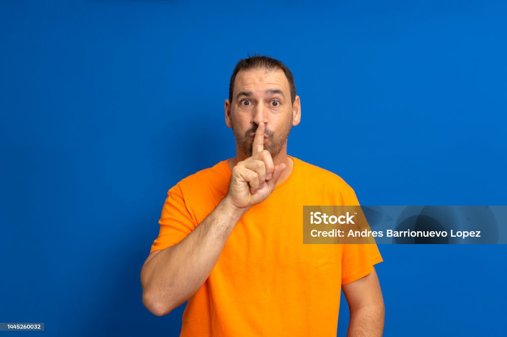Bearded latino man wearing orange casual t-shirt, pressing finger to lips as if asking to keep quiet, isolated over blue background. shhh gesture. Bearded latino man wearing orange casual t-shirt, pressing finger to lips as if asking to keep quiet, isolated over blue background. shhh gesture Adult Stock Photo