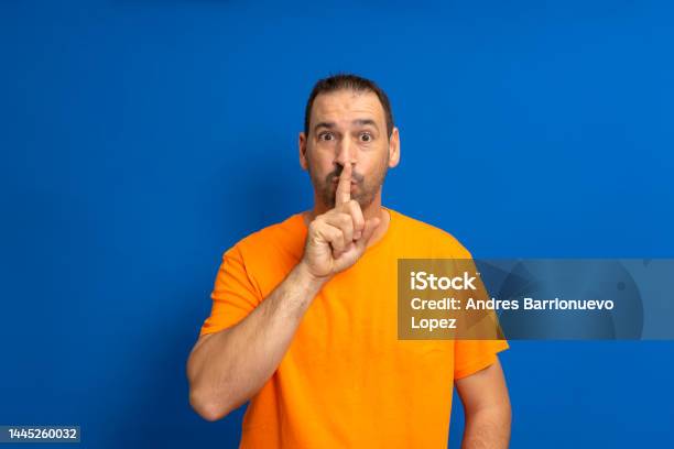 Bearded Latino Man Wearing Orange Casual Tshirt Pressing Finger To Lips As If Asking To Keep Quiet Isolated Over Blue Background Shhh Gesture Stock Photo - Download Image Now