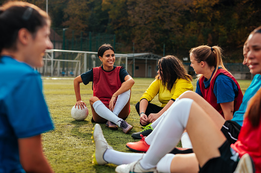A young female football team is sitting on the football field talking to each other, smiling and socializing.