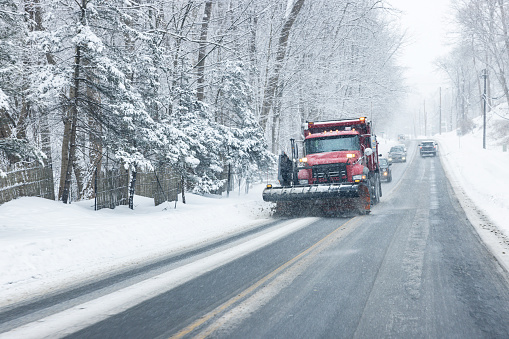 A large red town services snowplow dump truck is simultaneously plowing and spreading road salt; clearing and melting still falling snow and slush on the road surface as it lumbers uphill slowly on a steep, slippery, sloppy rural road hill near Rochester, NY during an early February western New York State blizzard storm.
