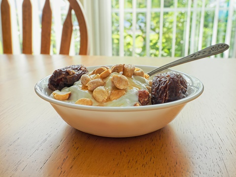 A healthy decadent snack of Greek yogurt with peanuts and pitted dates drizzled with honey with window and chair in background. The Deglet Nour dates are large and succulent in white bowl with spoon on hardwood table.