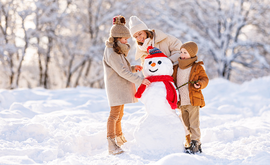 Young mom and two kids making snowman in winter park outdoors. Joyful little brother and sister in warm clothes happily helping mother decorate snow figure with carrot nose and hands with dry branches