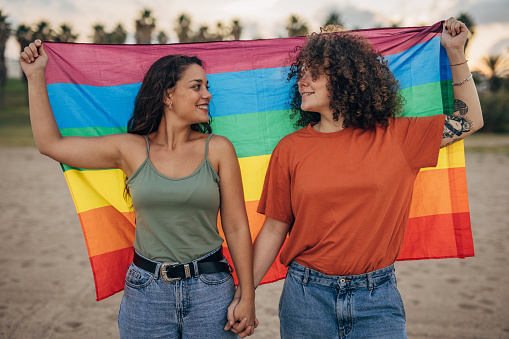 Two women, lesbian couple holding rainbow flag together outdoors on the beach.