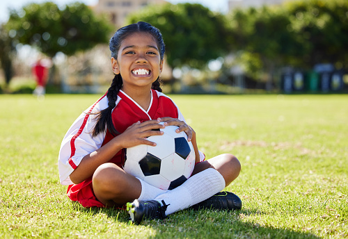 Sports, children and girl soccer player relax on grass with soccer ball, happy and excited at training. Fitness, smile and portrait of Indian child on field, ready for cardio, energy football workout