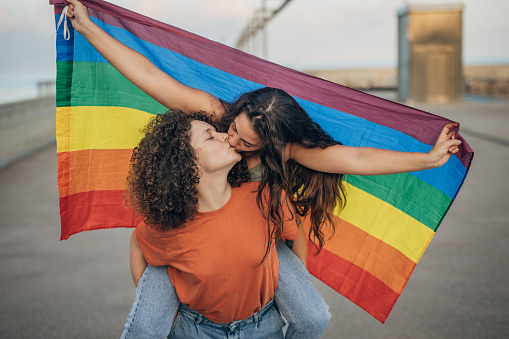 Two women, lesbian couple with rainbow flag kissing outdoors on the street.