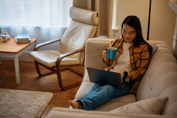 istock Smiling Asian woman drinking tea while using laptop on the sofa at home. 1445288693