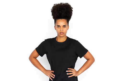 Portrait of confident young woman posing with hands on hips. African American woman wearing black T-shirt looking at camera against white background. Female beauty concept